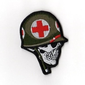 Puede incluir: Parche bordado con un casco militar con una cruz roja y una calavera. El casco es verde oliva con un acento rojo y costuras blancas. La calavera es blanca con cuencas de ojos y dientes negros.