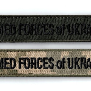 Puede incluir: Dos parches rectangulares bordados con el texto ARMED FORCES of UKRAINE en letras mayúsculas negras. El parche superior es de color verde oliva oscuro sólido, mientras que el inferior presenta un patrón de camuflaje digital beige y verde.