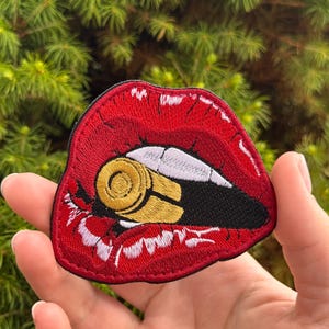 Peut inclure: Un écusson brodé représentant une paire de lèvres rouge vif avec des dents blanches mordant une douille de balle dorée. Le motif est souligné par un fil noir, créant une image graphique audacieuse avec un contraste marqué entre le rouge, l'or et le blanc.