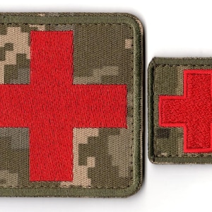 May include: Two embroidered patches featuring a red cross on a camouflage background. The larger patch is square, while the smaller one is rectangular. The patches have a green border.