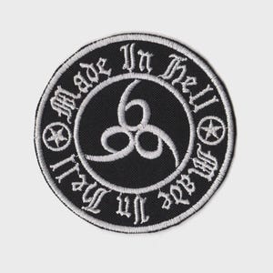 May include: A circular black embroidered patch featuring white stitching. The center displays a stylized triple-six symbol. The border contains the text Made In Hell in a gothic font, separated by two small five-pointed stars on each side.