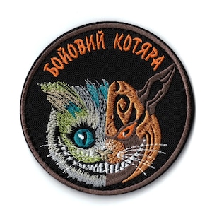 以下が含まれることがあります： 刺繍パッチ、顔が分かれた猫のデザイン。片側は緑と青、もう片側は茶色とオレンジ色です。パッチには、猫の頭の上にアーチ状にオレンジ色で「БОЙОВИЙ КОТЯРА」という文字が黒い背景に書かれています。