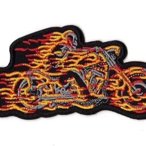 May include: An embroidered patch featuring a skeleton riding a motorcycle engulfed in bright yellow, orange, and red flames. The design is set against a black background with a thick black border, showcasing detailed grey stitching for the bike.