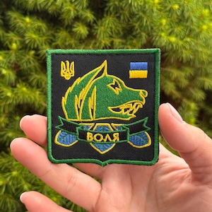 May include: A square embroidered patch with a black background and a green border. It features a green wolf head, a yellow Ukrainian trident, a small blue and yellow flag, and a black banner with the yellow Cyrillic text "ВОЛЯ" above a blue shield.