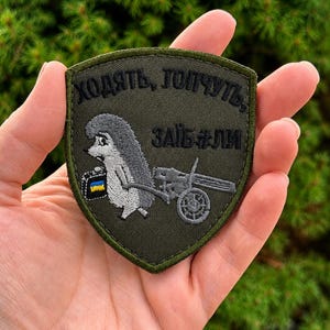 May include: A shield-shaped olive green embroidered patch featuring a cartoon hedgehog carrying a small black case with a blue and yellow flag design and pulling a grey machine gun. The Cyrillic text "ХОДЯТЬ, ТОПЧУТЬ, ЗАЇБАЛИ" is stitched in black thread at the top.