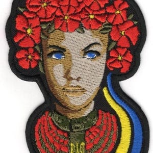 May include: Embroidered patch featuring a woman with blue eyes and a floral crown. The patch includes a red and green outfit, and a blue and yellow ribbon. The patch has a black border.