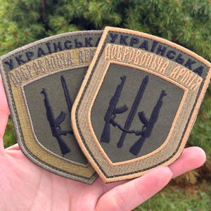 Puede incluir: Dos parches bordados en forma de escudo con fondo verde oliva y borde negro. El diseño incluye dos rifles negros dispuestos verticalmente y el texto en cirílico "УКРАЇНСЬКА ДОБРОВОЛЬЧА АРМІЯ" bordado en hilo negro en el borde superior.