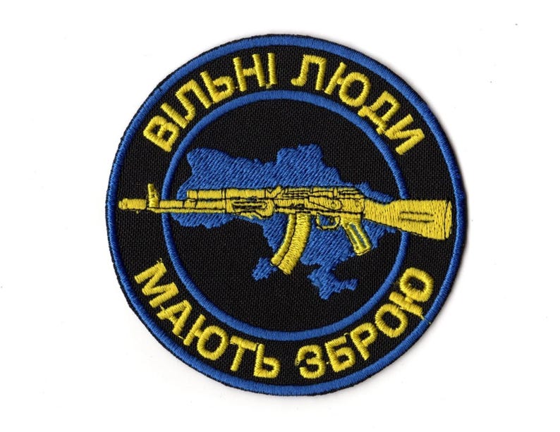 May include: Embroidered patch with a black background and a blue and yellow design. The patch features a blue outline of Ukraine, a yellow AK-47 rifle, and yellow text in a circular pattern that reads "Free People Have Weapons."