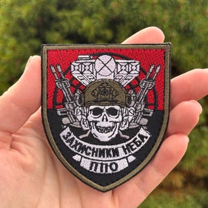 Op de afbeelding: Een schildvormige geborduurde patch met een schedel die een helm en headset draagt, geflankeerd door twee geweren. Boven de schedel bevindt zich een radarantenne. De achtergrond is verdeeld in rood en zwart. Witte tekst "ЗАХИСНИКИ НЕБА" en "ППО".