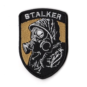 Op de afbeelding: Zwart en goudkleurig embleem met het woord "STALKER" in wit geborduurd. Het ontwerp toont een persoon met een gasmasker en beschermende kleding, geschikt voor kleding.