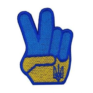 Pode incluir: Remendo bordado em forma de mão fazendo o sinal de paz. Os dedos são azuis e a palma é amarela. Um contorno azul emoldura o desenho. Um tridente amarelo é bordado na palma.