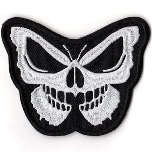 May include: A black embroidered patch in the shape of a butterfly with a skull design. The skull is white with detailed stitching, featuring eye sockets, a nose, and teeth. The patch has a black border.