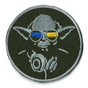 May include: A circular embroidered patch featuring a gray character with pointed ears wearing glasses with blue and yellow lenses. The character is depicted with a pair of headphones around the neck, all set against a dark olive green background.