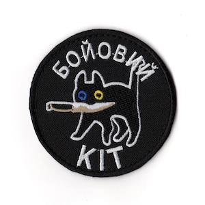 May include: A round, black embroidered patch featuring a cartoon cat with a knife. The cat has blue and yellow eyes and is outlined in white. The patch has white text that reads "БОЙОВИЙ KIT".