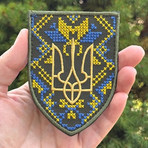Könnte beinhalten: Ein schildförmiger, bestickter Aufnäher mit dunkelolivgrünem Hintergrund und einem zentralen gelben ukrainischen Dreizack-Symbol. Das Design zeigt komplexe Kreuzstichmuster in leuchtendem Gelb und Blau, eingefasst von einem dunkelgrünen Rand.