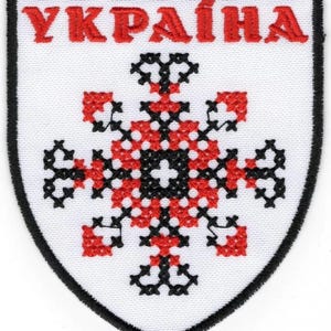 Puede incluir: Parche blanco con borde negro y la palabra ucraniana "УКРАЇНА" en letras rojas. El diseño central es un patrón floral o de copo de nieve estilizado en rojo y negro sobre fondo blanco.