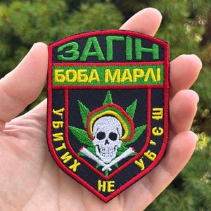 May include: A shield-shaped embroidered patch with a black background and red border. It features a white skull with a green, yellow, and red halo, centred over green cannabis leaves and two crossed white bones. Text in Cyrillic reads ЗАГІН БОБА МАРЛІ.