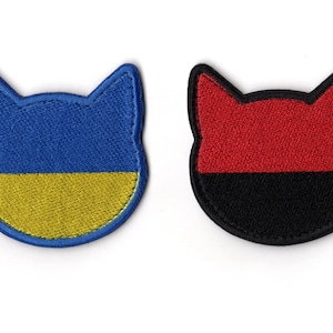 Puede incluir: Dos parches bordados con forma de cabeza de gato. Uno presenta un diseño azul y amarillo, mientras que el otro tiene una combinación de colores rojo y negro. Ambos parches tienen un borde cosido.