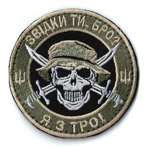 May include: Embroidered patch featuring a skull wearing a camouflage hat, with crossed swords behind it. The patch has text in a circular pattern around the skull, with a black background and olive green border.