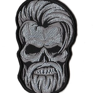 May include: An embroidered patch featuring a stylized skull with a thick beard and a pompadour hairstyle. The design is rendered in shades of grey and silver thread against a black background, with a black stitched border outlining the shape.