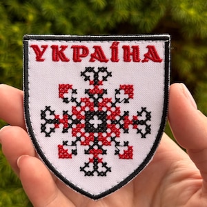 May include: A shield-shaped embroidered patch with a white background and a black border. The text "УКРАЇНА" is stitched in red at the top. Below the text is a symmetrical geometric cross pattern created with red and black cross-stitching.