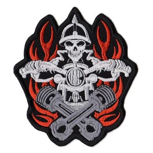May include: An embroidered patch featuring a white skull wearing a helmet, positioned above two crossed grey engine pistons. The design is set against a black background with vibrant red flame accents surrounding the central motorcycle-themed imagery.