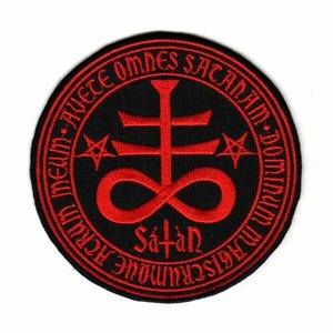 May include: A round black patch with red embroidered text and symbols. The design includes a cross, an infinity symbol, stars, and the word "Satan". The outer ring of the patch features additional text.