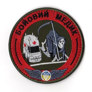 Pode incluir: Patch bordado circular com borda vermelha e fundo verde. O design apresenta uma ambulância, uma figura da Morte e o texto "БОЙОВИЙ МЕДИК". O patch também inclui uma bandeira ucraniana e um par de asas na parte inferior.