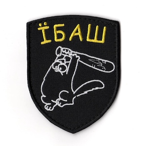 Puede incluir: Parche bordado negro con forma de escudo. El parche presenta un gato de dibujos animados sosteniendo un bate de béisbol, con el texto amarillo "ЇБАШ" encima del gato. El gato y el bate están delineados en blanco.