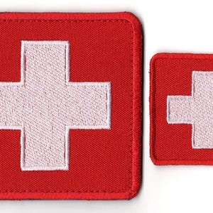 Op de afbeelding: Twee rode vierkante patches met een wit kruis in het midden, die lijken op de Zwitserse vlag. De patches zijn van verschillende formaten, met een gestikte rand. De grotere patch staat links en de kleinere rechts.