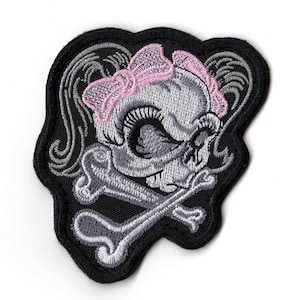 Puede incluir: Parche bordado con una calavera gris con un lazo rosa, huesos cruzados grises y remolinos grises decorativos sobre un fondo negro. El parche tiene un borde negro, adecuado para ropa o accesorios.