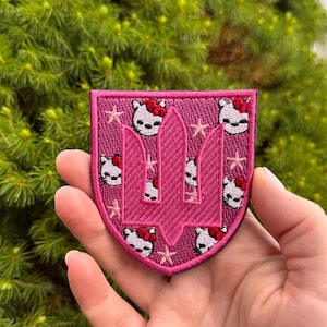 May include: A shield-shaped embroidered patch in shades of pink and magenta. The design features a central dark pink trident symbol surrounded by a repeating pattern of white cat faces with red bows and small light pink stars on a magenta background.