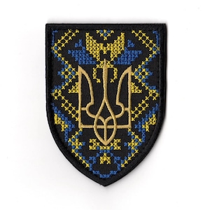 Puede incluir: Un parche bordado negro con forma de escudo. Presenta un emblema de tridente dorado, rodeado de patrones de punto de cruz azules y amarillos. El borde es negro.