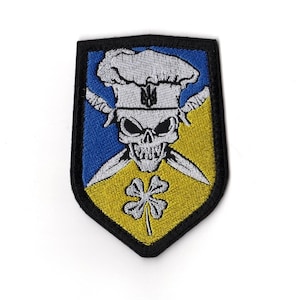 Könnte beinhalten: Gestickter Aufnäher in Schildform. Das Design zeigt einen Totenkopf mit Kochmütze, gekreuzten Messern und einem vierblättrigen Kleeblatt. Der Hintergrund ist in blaue und gelbe Abschnitte unterteilt, mit einem schwarzen Rand.