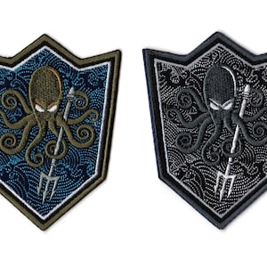 Puede incluir: Dos parches bordados con forma de escudo. Uno presenta un pulpo verde oliva con un tridente sobre un fondo azul estampado, el otro un pulpo gris sobre un fondo negro estampado.