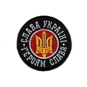 Puede incluir: Un parche redondo bordado en negro con texto blanco en círculo. El texto dice "¡Gloria a Ucrania! ¡Gloria a los héroes!" Un círculo rojo en el centro presenta un emblema de tridente dorado.