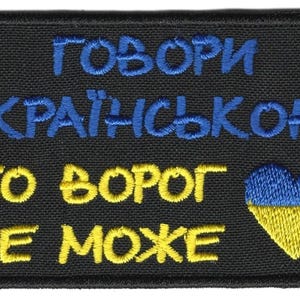Puede incluir: Parche bordado negro con texto azul y amarillo en ucraniano. El texto dice "¡Habla ucraniano! Porque el enemigo no puede." Un diseño en forma de corazón azul y amarillo está en el lado derecho.