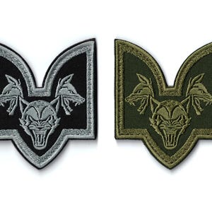 Puede incluir: Dos parches bordados. Uno es negro con detalles plateados, el otro es verde oliva con detalles caqui. Ambos presentan un diseño estilizado de una bestia gruñona con dos cabezas de lobo que flanquean la cara central.