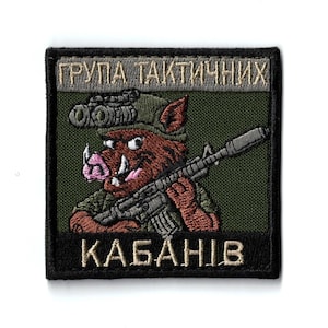 May include: Embroidered patch featuring a cartoon boar in military attire, holding a rifle. The patch has text in a foreign language at the top and bottom. The boar is brown with a pink snout and tusks, wearing a green hat and uniform. The background is green.
