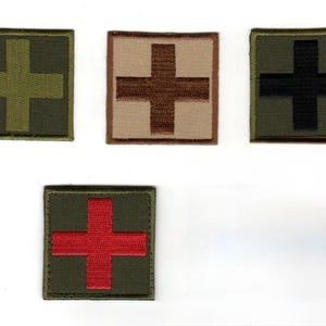May include: Four square embroidered patches featuring a central cross design. One patch is olive green with a lighter green cross, one is tan with a brown cross, one is olive green with a black cross, and one is olive green with a red cross.