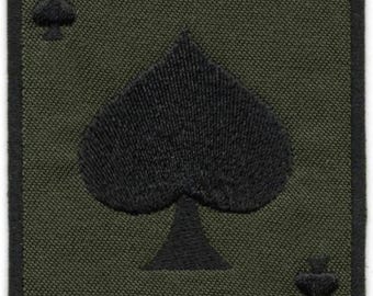May include: A rectangular embroidered patch featuring an olive green background with a black border. The design depicts an ace of spades playing card, with a large black spade symbol in the center and the letter A with a small spade in each corner.