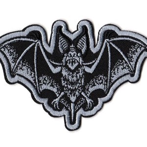 Peut inclure: Un écusson brodé représentant une chauve-souris aux ailes déployées. Le motif utilise du fil noir et gris clair pour créer une illustration détaillée de la créature, avec une bordure épaisse cousue en gris clair tout autour de la forme.