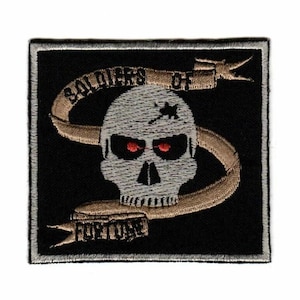May include: A square embroidered patch with a black background featuring a light gray skull with red eyes and a dark mark on the forehead. A tan ribbon wraps around the skull with the text Soldiers of Fortune embroidered in black.