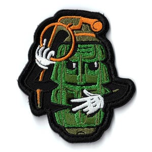 Puede incluir: Parche bordado con una granada verde de estilo caricatura con brazos negros y un borde negro. La granada sostiene un lazo con una mano enguantada y hace un signo de la paz con otra mano enguantada.
