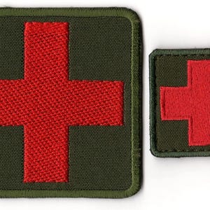 Può includere: Due toppe ricamate con una croce rossa su sfondo verde scuro. La toppa più grande è quadrata, quella più piccola rettangolare. Entrambe hanno un bordo verde.