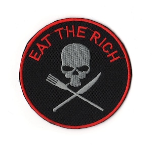 Peut inclure: Écusson brodé rond, fond noir, bordure rouge. Il présente un crâne gris, une fourchette et un couteau croisés, et le texte rouge "EAT THE RICH".