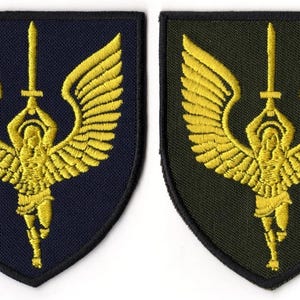 Puede incluir: Dos parches bordados con una figura alada dorada sosteniendo una espada. Uno tiene un fondo azul marino, el otro verde oliva. La figura y las alas están ricamente bordadas. Los parches tienen forma de escudo.