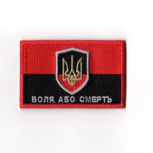 Puede incluir: Parche rectangular con parte superior roja e inferior negra. Un escudo blanco con un emblema de tridente dorado está centrado. El texto "ВОЛЯ АБО СМЕРТЬ" está bordado en blanco en la sección negra.