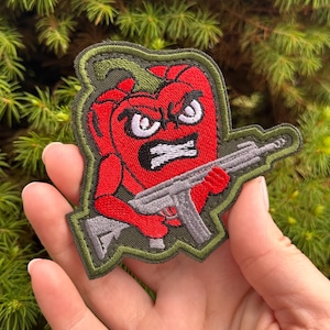 Könnte beinhalten: Ein gestickter Aufnäher, der eine rote Paprika-Figur mit wütendem Gesichtsausdruck, weißen Augen und schwarzem Mund zeigt. Die Figur hält ein graues Gewehr. Der Aufnäher hat einen olivgrünen Rand und einen passenden olivgrünen Hintergrund.