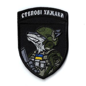 Könnte beinhalten: Schwarzer, gestickter Aufnäher mit einem wolfsähnlichen Charakter in Militäruniform. Der Aufnäher enthält den Text "СТЕПОВІ ХИЖАКИ" in Weiß, mit einem Detail der ukrainischen Flagge. Das Design verwendet Grün-, Grau- und Schwarztöne.